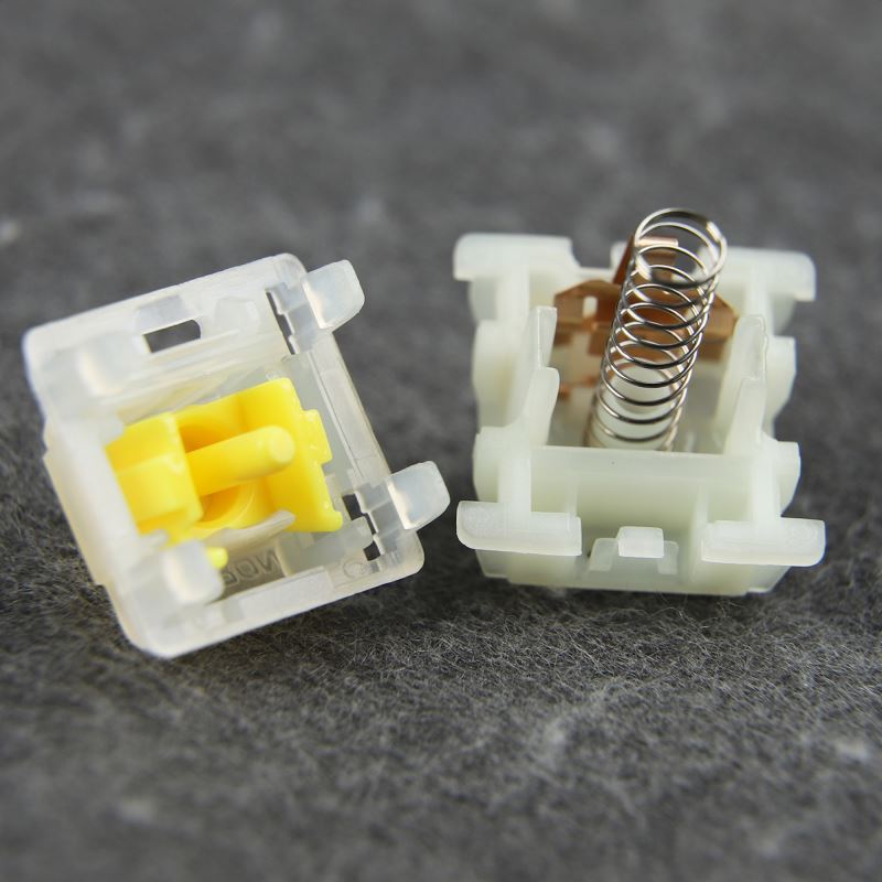 A close-up image of two disassembled Gateron Milky Yellow Pro switches. The switches are transparent white with a yellow stem and spring. The housing is open, showing the internal components.