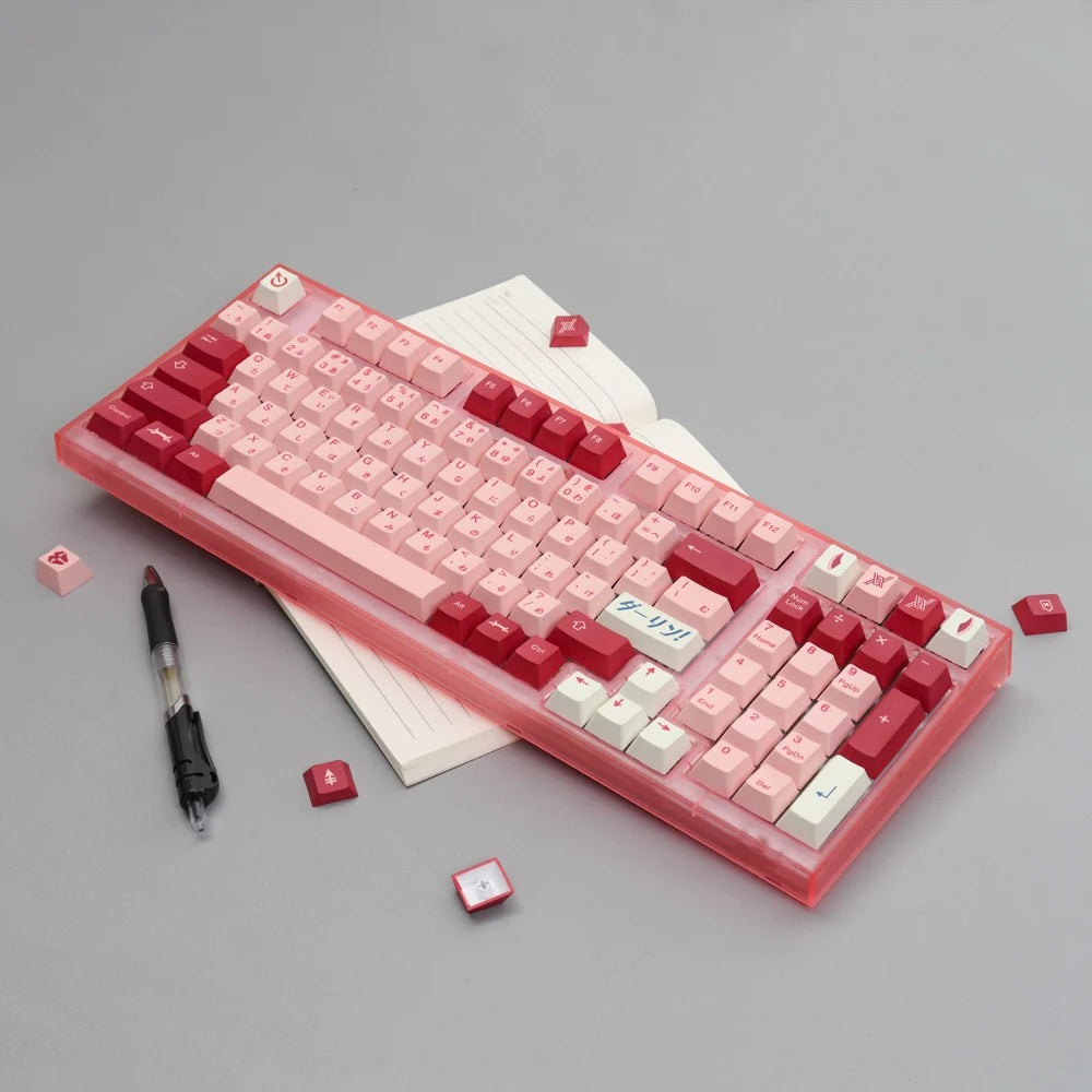 A mechanical keyboard with pink and red keycaps, featuring a celestial theme. The keyboard is placed on a desk with a notebook and pen nearby.