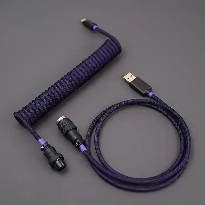 A coiled keyboard cable with a purple braided outer sheath and gold-colored connectors. The cable features a USB-C connector on one end and an aviator connector on the other.