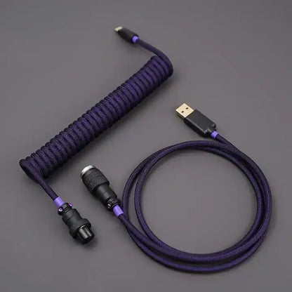 A coiled keyboard cable with a purple braided outer sheath and gold-colored connectors. The cable features a USB-C connector on one end and an aviator connector on the other.